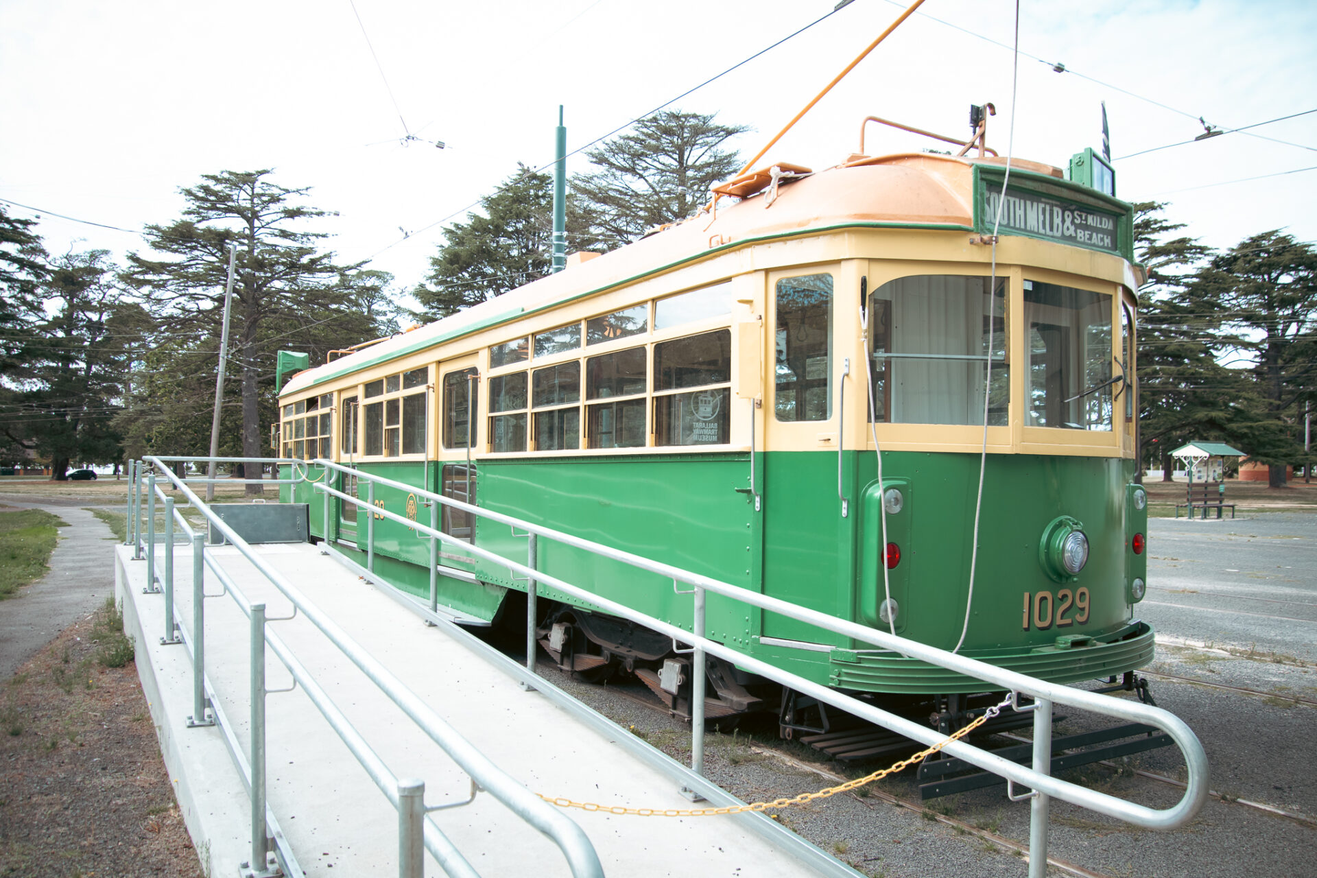 Photograph of a vintage green and cream ex Melbourne tram numbered 1029 parked on tracks beside a metal ramp that provides wheel chair access to the tram interior.