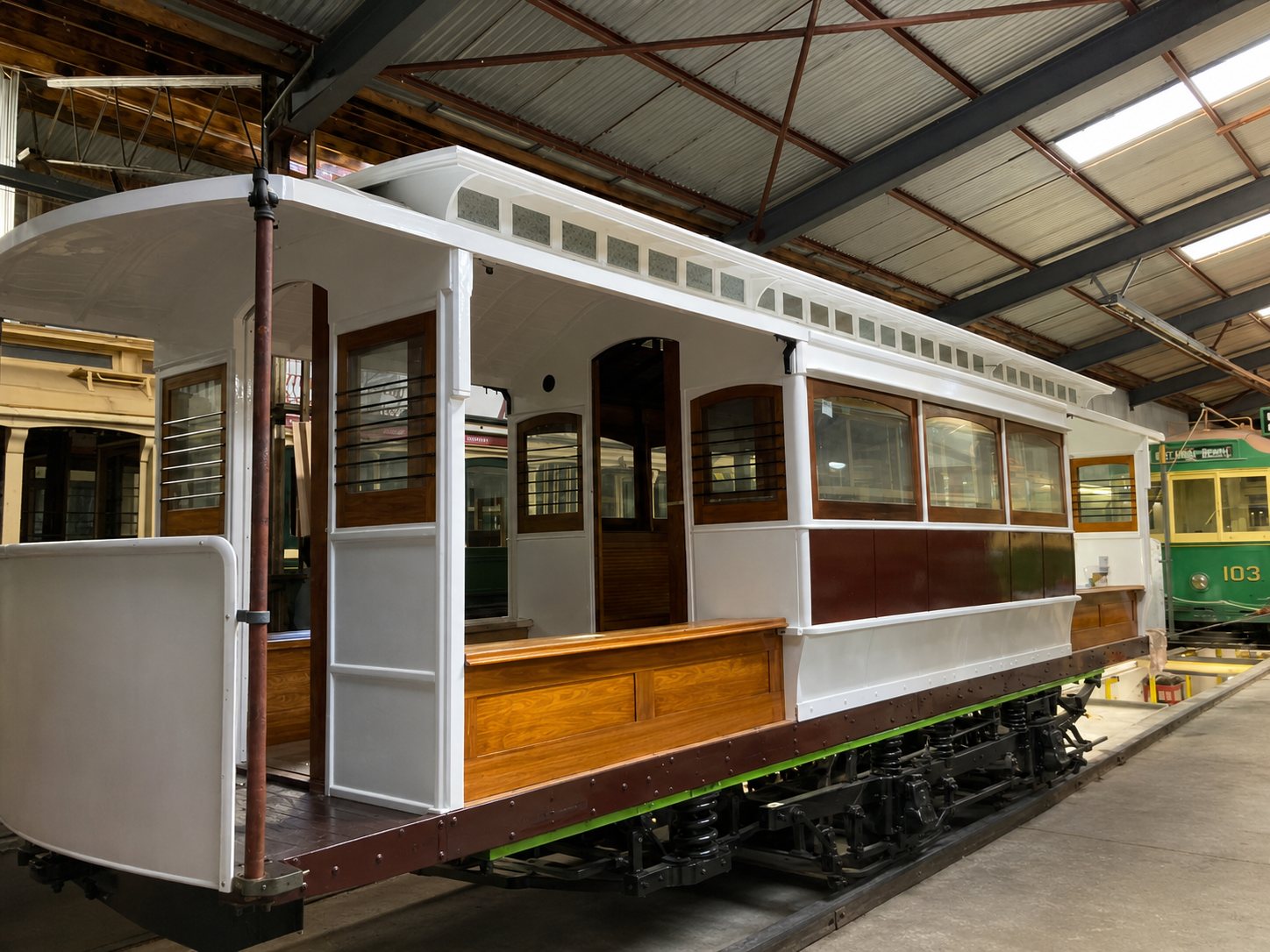 A white and brown tram with wooden benches sits inside a tram depot with a corrugated metal roof. The tram has large windows and an open-air platform at the front.