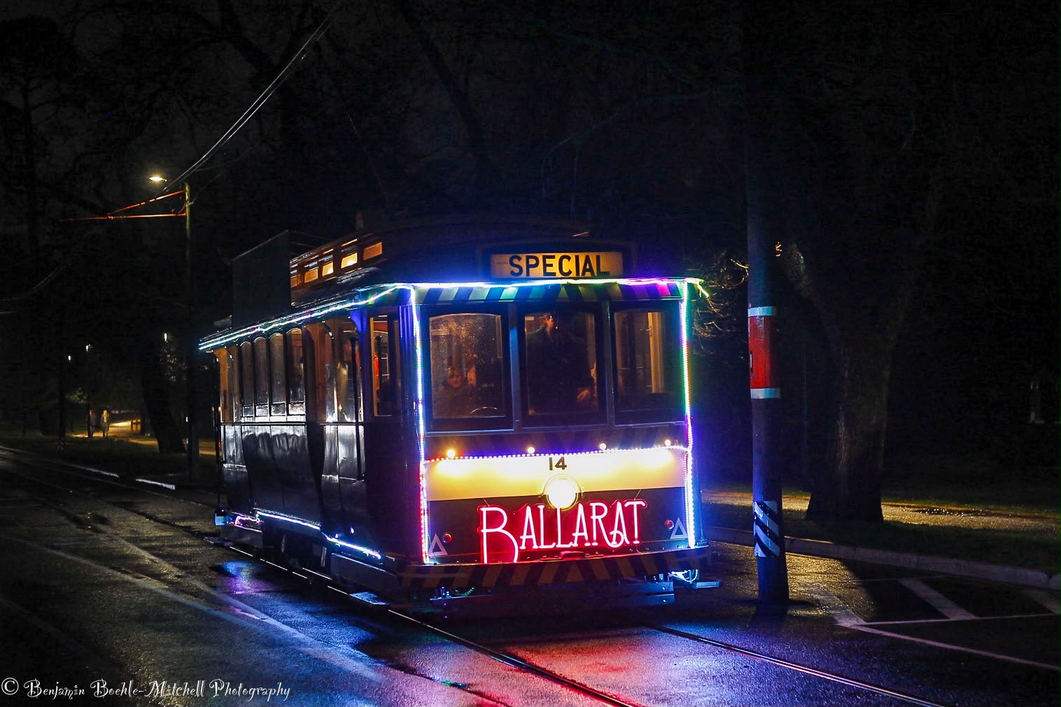 A decorated tram labeled "BALLARAT" and "SPECIAL" travels on tracks at night, illuminated by colorful festive lights and reflections on the wet ground.