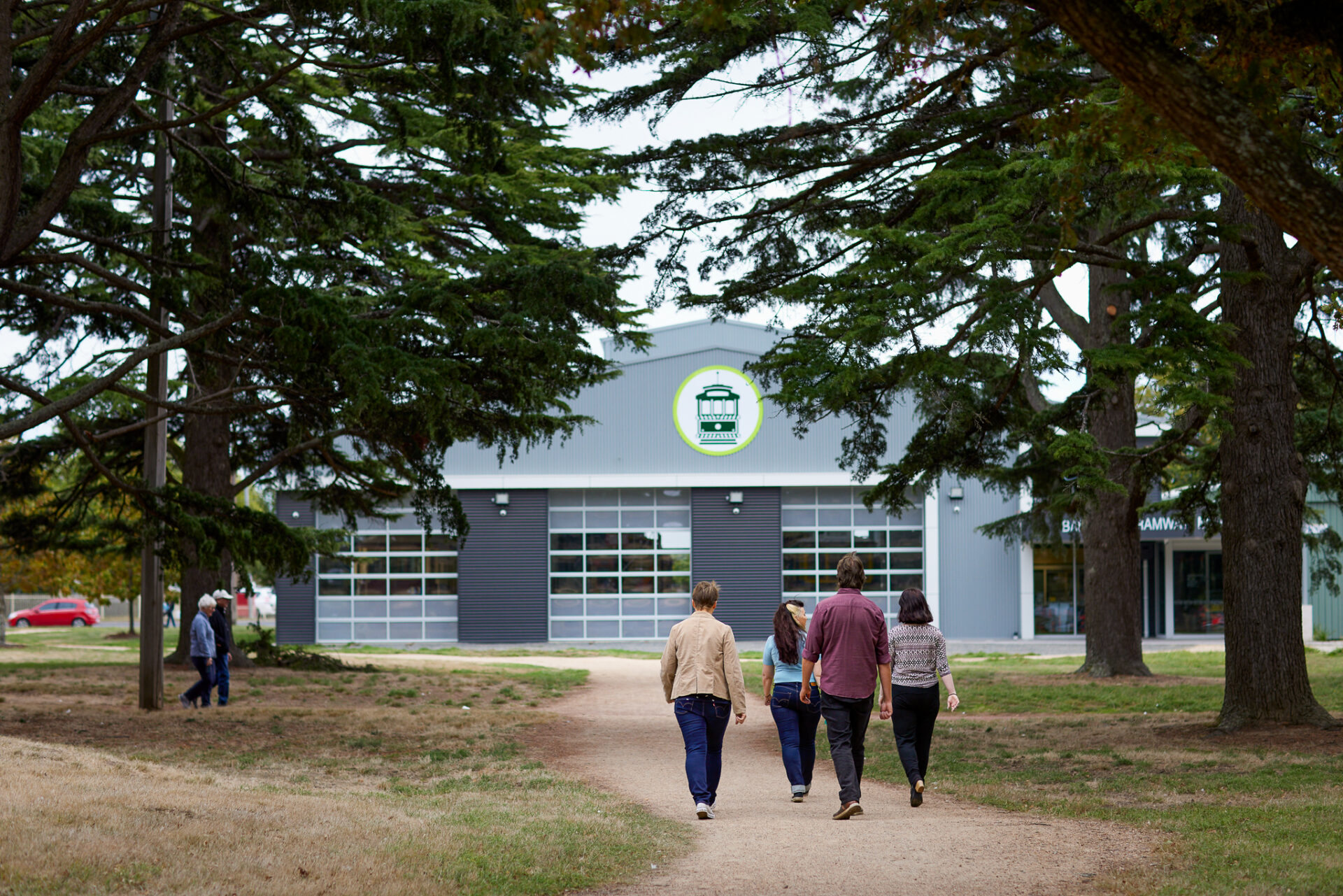 Four people walk on a path toward our modern tramcar depot building with large doors to allow for tramcars, featuring a prominent green trolley logo above the entrance, framed by large trees.