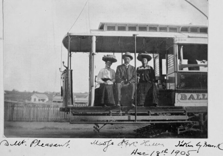 A black and white photograph of three people seated on an open-air part of an early tramcar. The tram has ornate trim and the word "BALL" is visible on its side. The people are dressed in early 20th-century attire, with the man wearing hats and the women wearing wide-brimmed hats. There are buildings and a fence visible in the background.