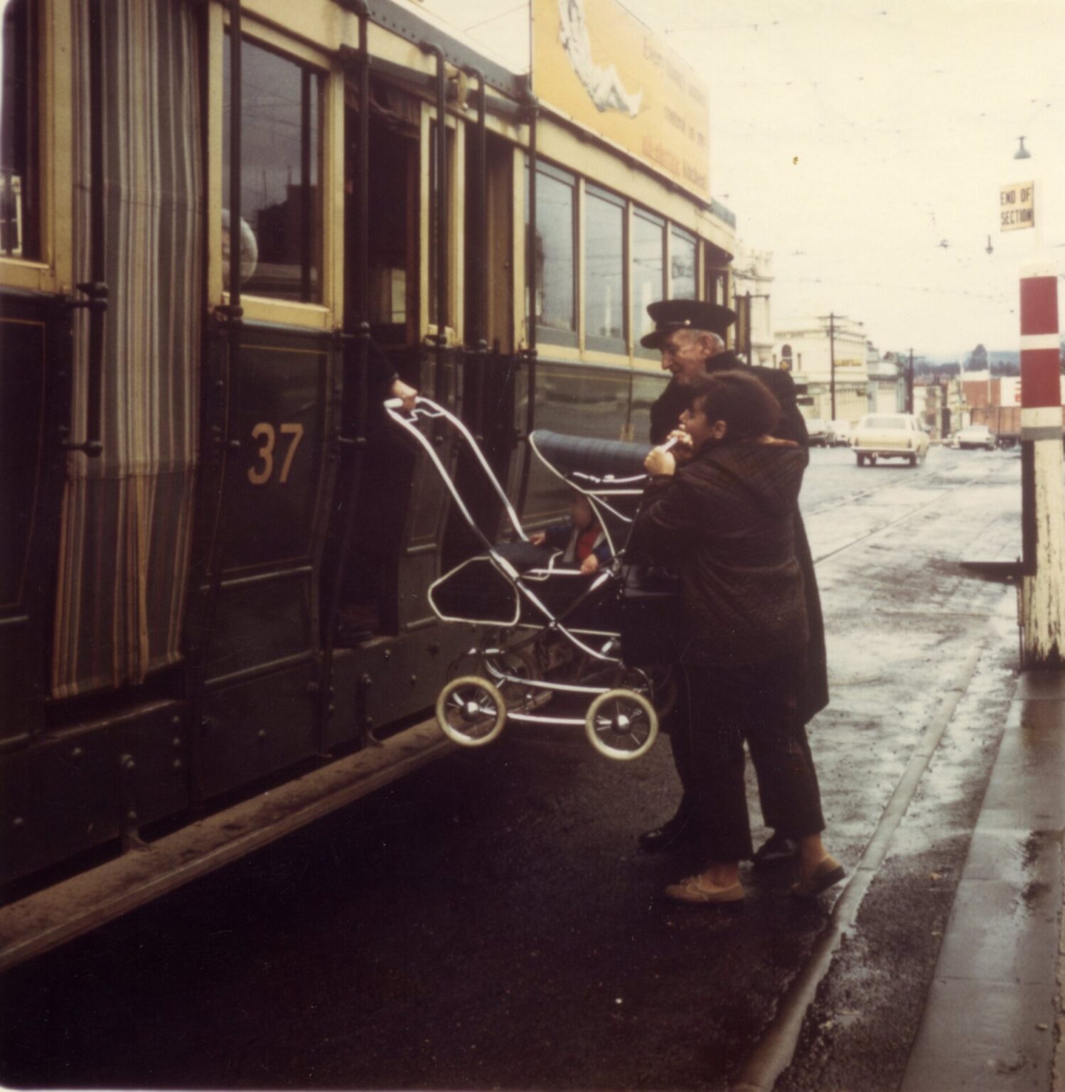 Photograph showing a person with a baby stroller boarding a vintage tram numbered 37, assisted by a uniformed conductor. The scene captures a wet urban street with visible tram tracks, a "End of Section" sign, and a few cars in the background, highlighting public transportation and accessibility.